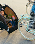 A child rides in a Burley trailer behind a woman riding a Bluejay e-bike in Mint Green 