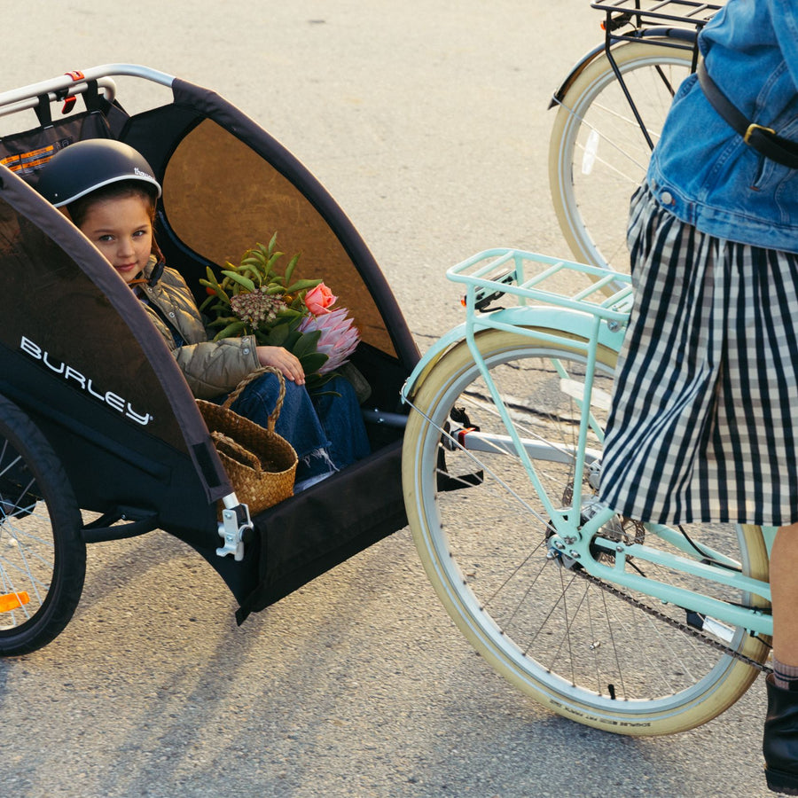 A child rides in a Burley trailer behind a woman riding a Bluejay e-bike in Mint Green 