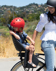 A woman smiles at her child while sitting on a Bluejay e-bike in Classic Black with a rear child seat. 