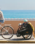 Woman riding Bluejay e-bike with two kids in a yellow Burley trailer.