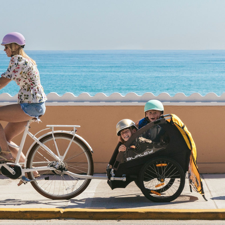 Woman riding Bluejay e-bike with two kids in a yellow Burley trailer.